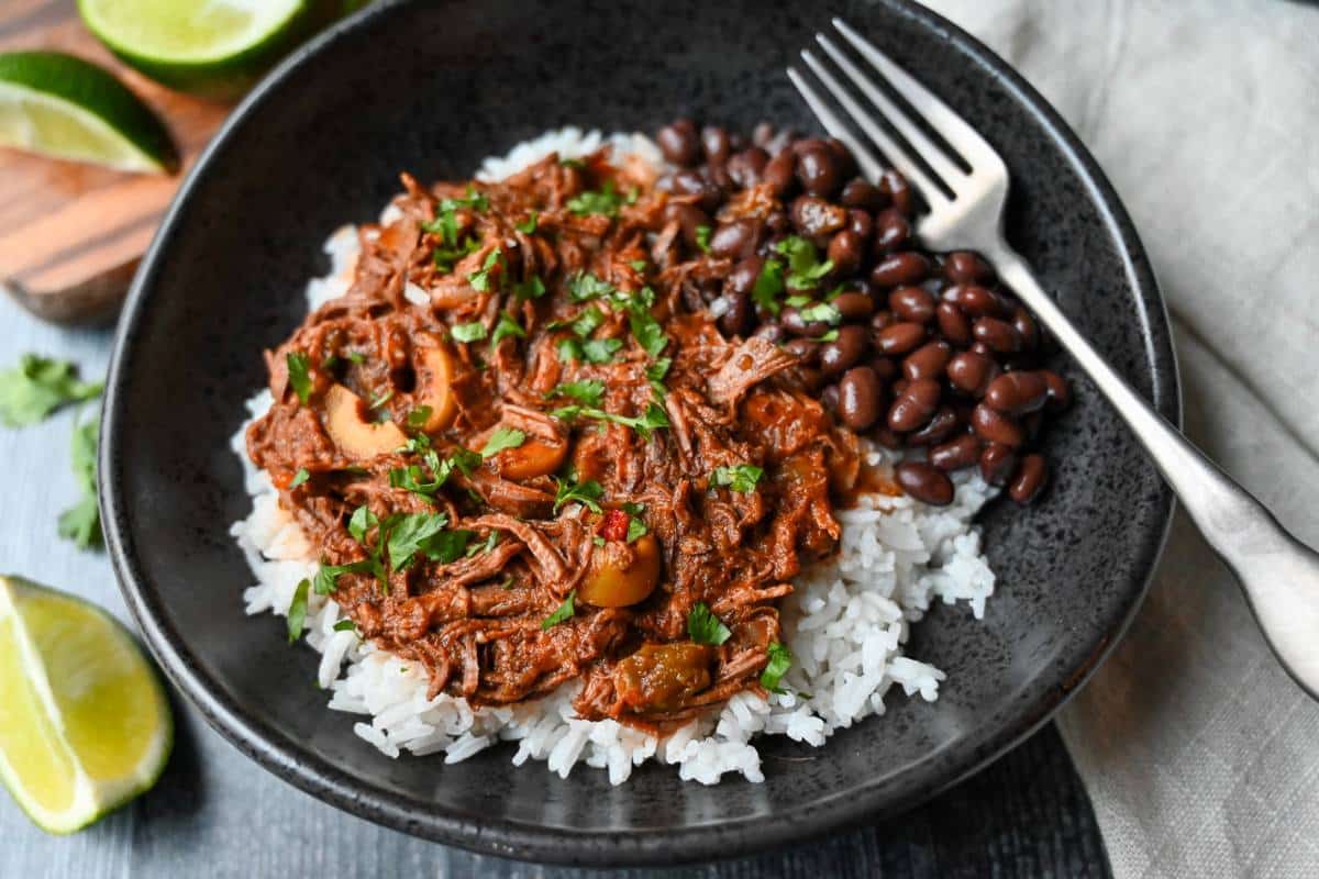 a bowl of shredded cuban beef over rice with cilantro and lime wedges