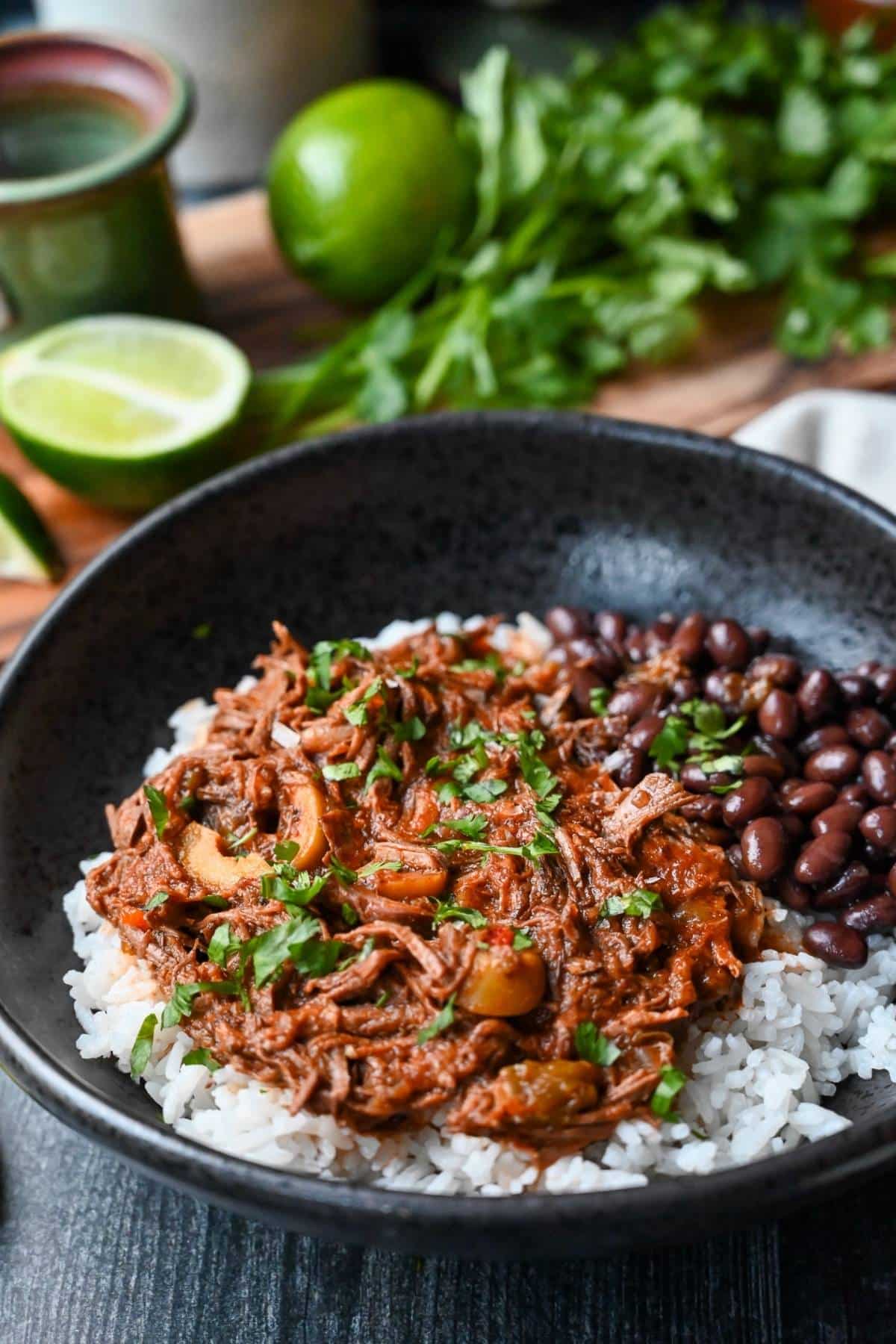 a bowl of cuban beef with rice and beans with limes and a bunch of cilantro behind it