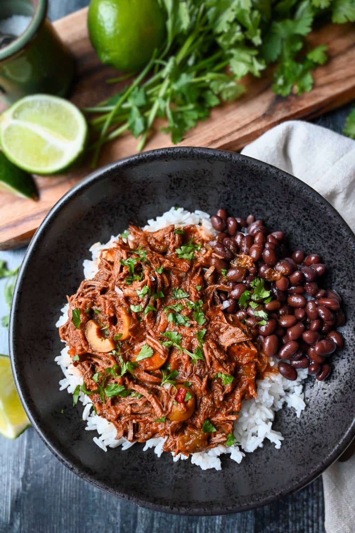a bowl of cuban shredded beef over rice with black beans and a cutting board with limes and herbs behind it