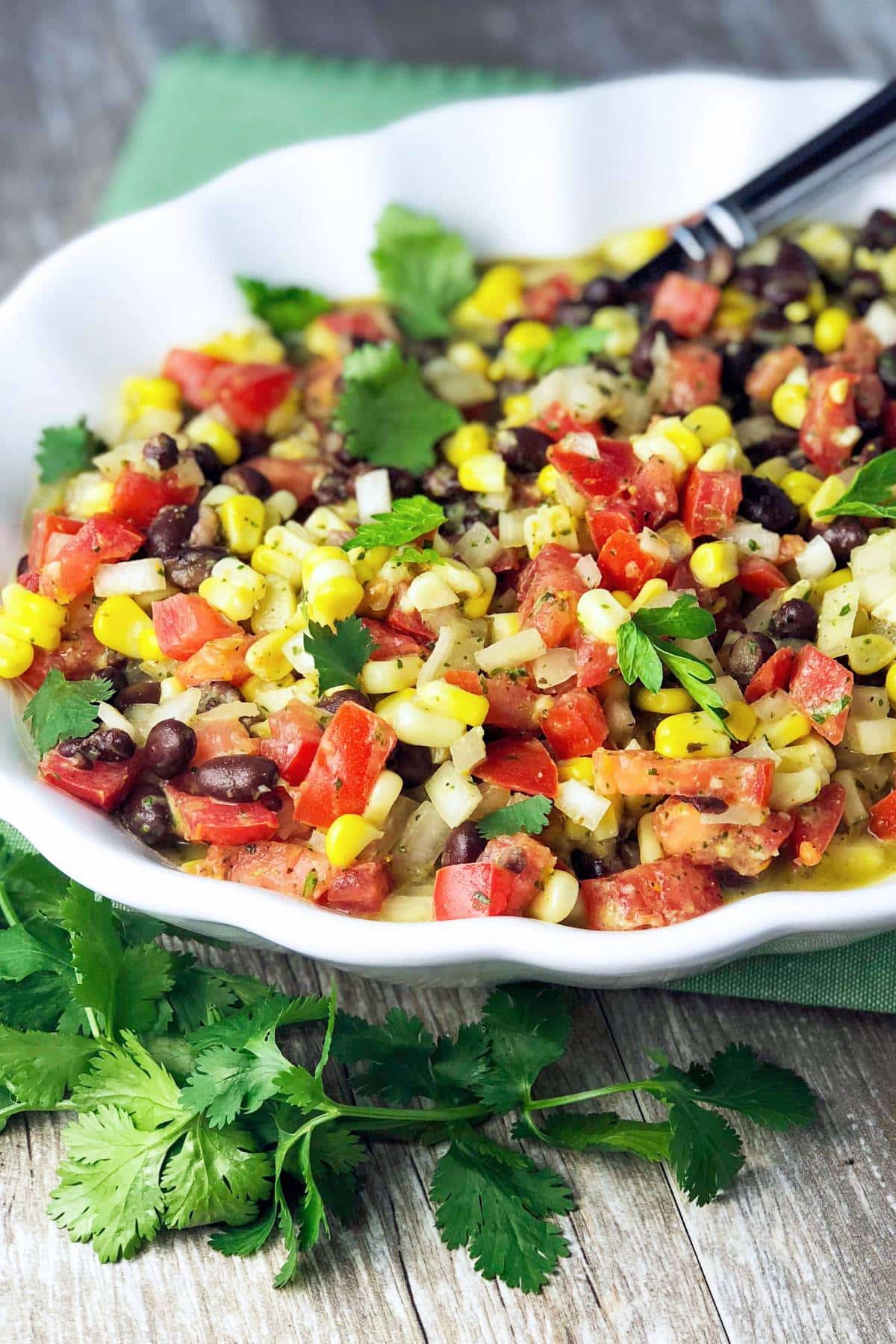 fiesta corn and bean salad in a white serving bowl with a spoon and a bunch of cilantro on the side