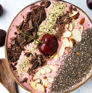 close up of a cherry smoothie bowl in a coconut shell topped with almonds, hemp seeds, dark chocolate and chia seeds