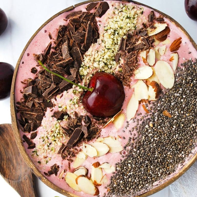 close up of a cherry smoothie bowl in a coconut shell topped with almonds, hemp seeds, dark chocolate and chia seeds