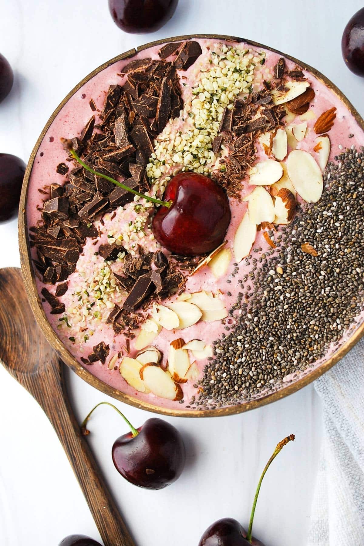 overhead photo of a cherry smoothie bowl topped with chia seeds, almonds, dark chocolate, and hemp seeds with fresh cherries