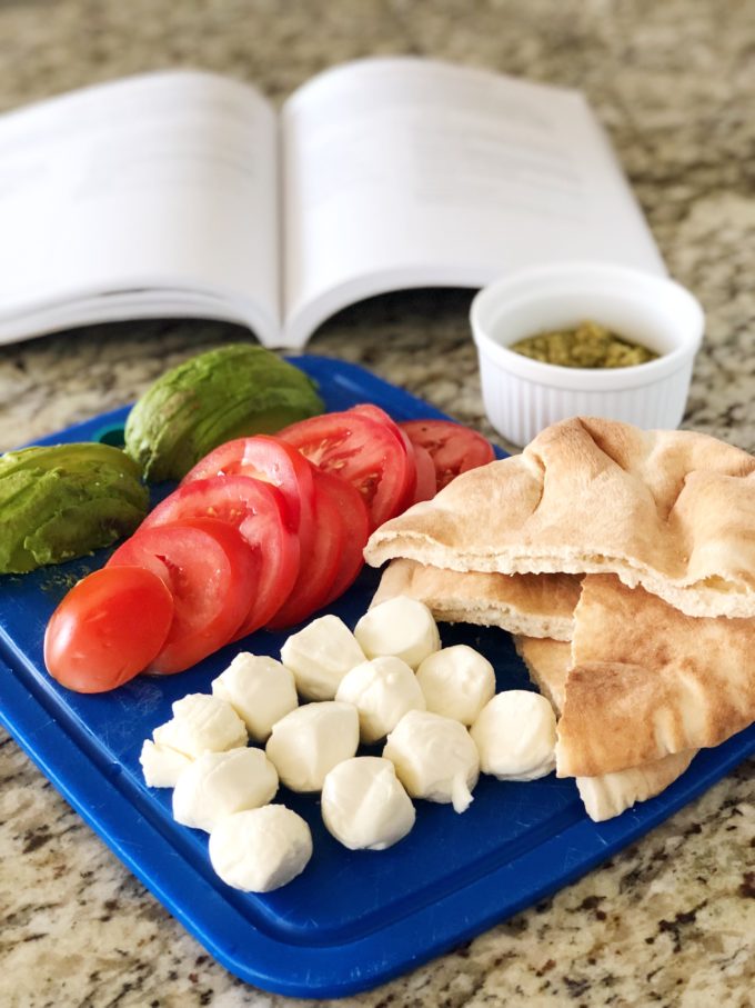 Cutting board with mozzarella balls, sliced pitas, sliced tomatoes, sliced avocados, and olive oil in a ramekin