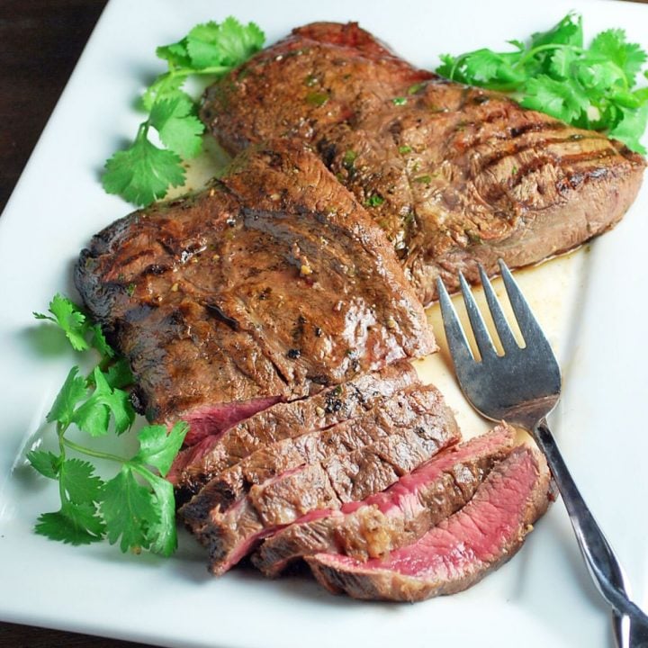 two flat iron steaks on a platter with half a steak sliced for serving. Fresh parsley and lime garnish the platter.