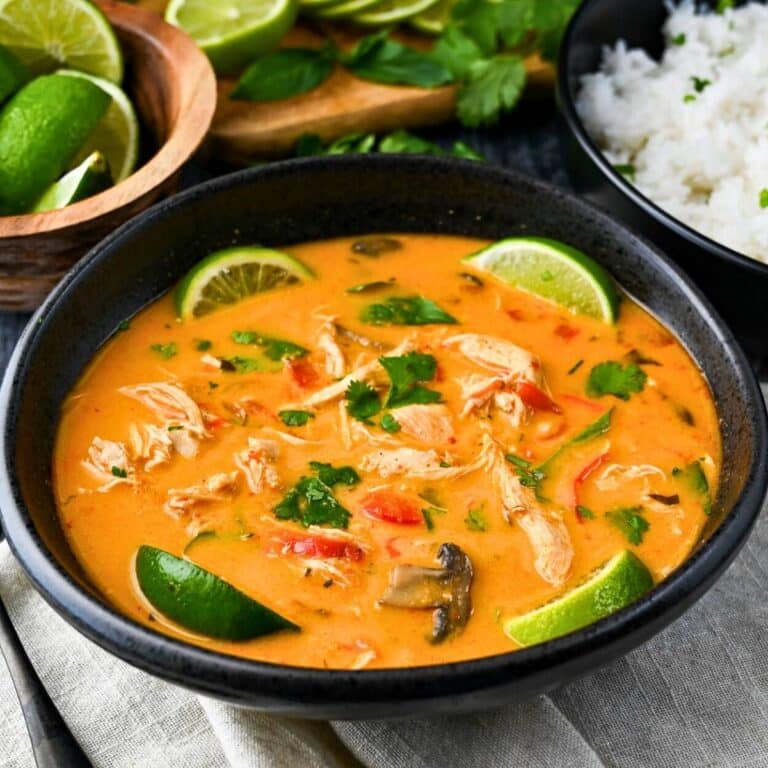 side view of a bowl of red curry chicken soup with fresh cilantro and lime wedges