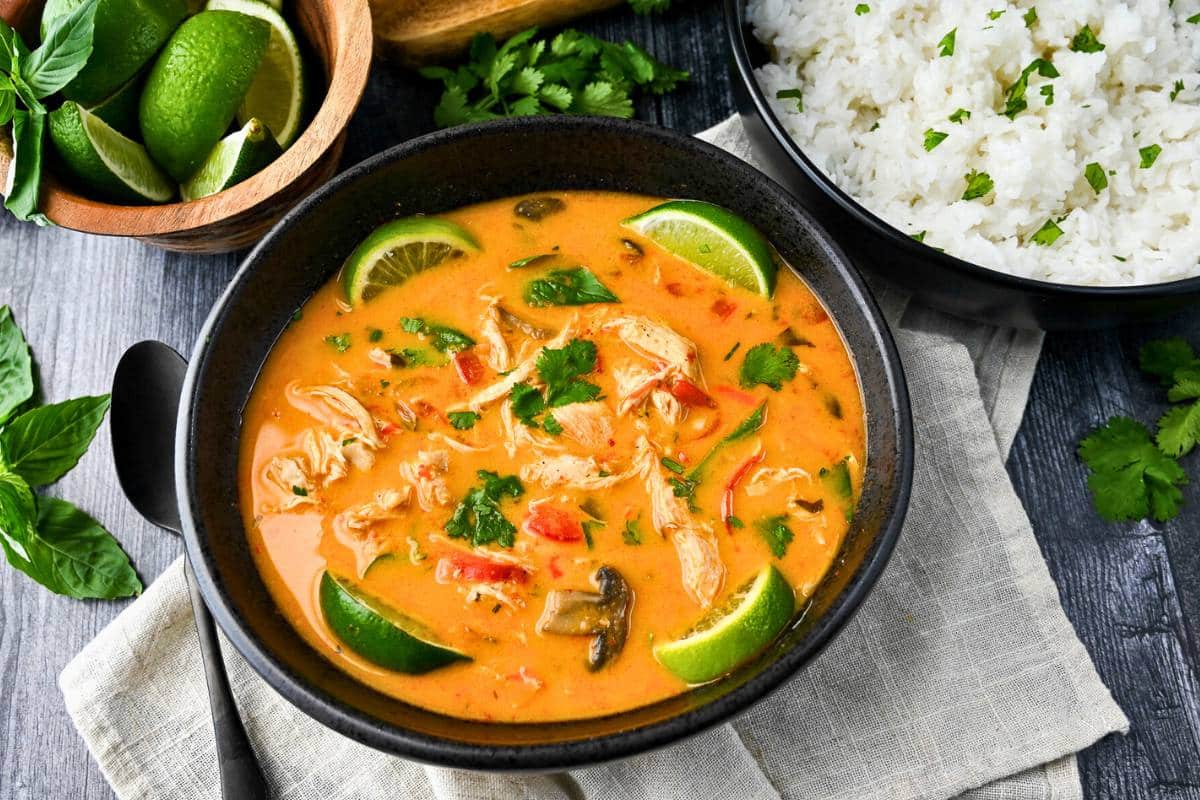 a bowl of chicken curry soup with a bowl of rice and limes next to it
