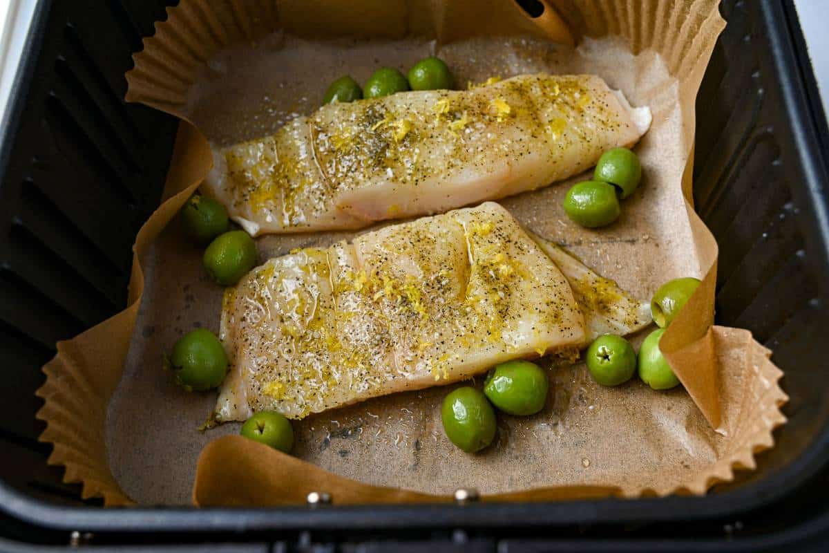 two halibut fillets in an air fryer basket with olives and a parchment liner