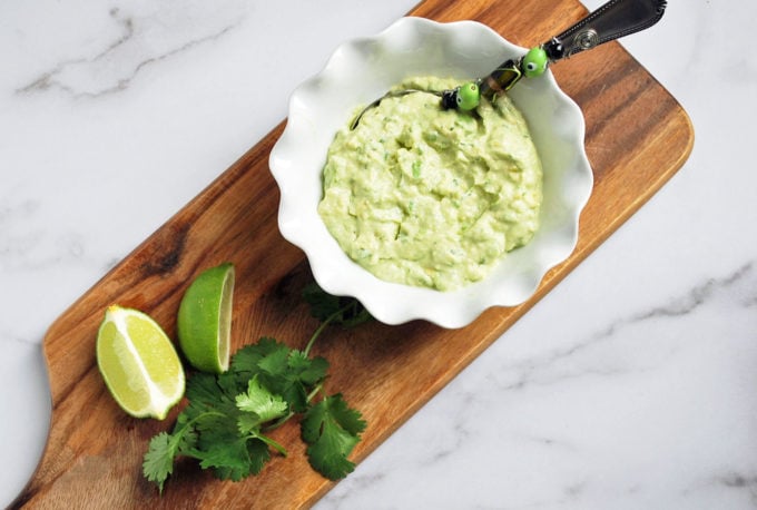 overhead photo of bowl of avocado dip on a wood board with lime slices and cilantro