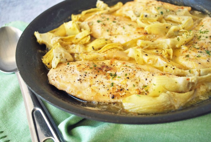 A close up of chicken breasts with artichokes and lemon in a skillet with a green napkin