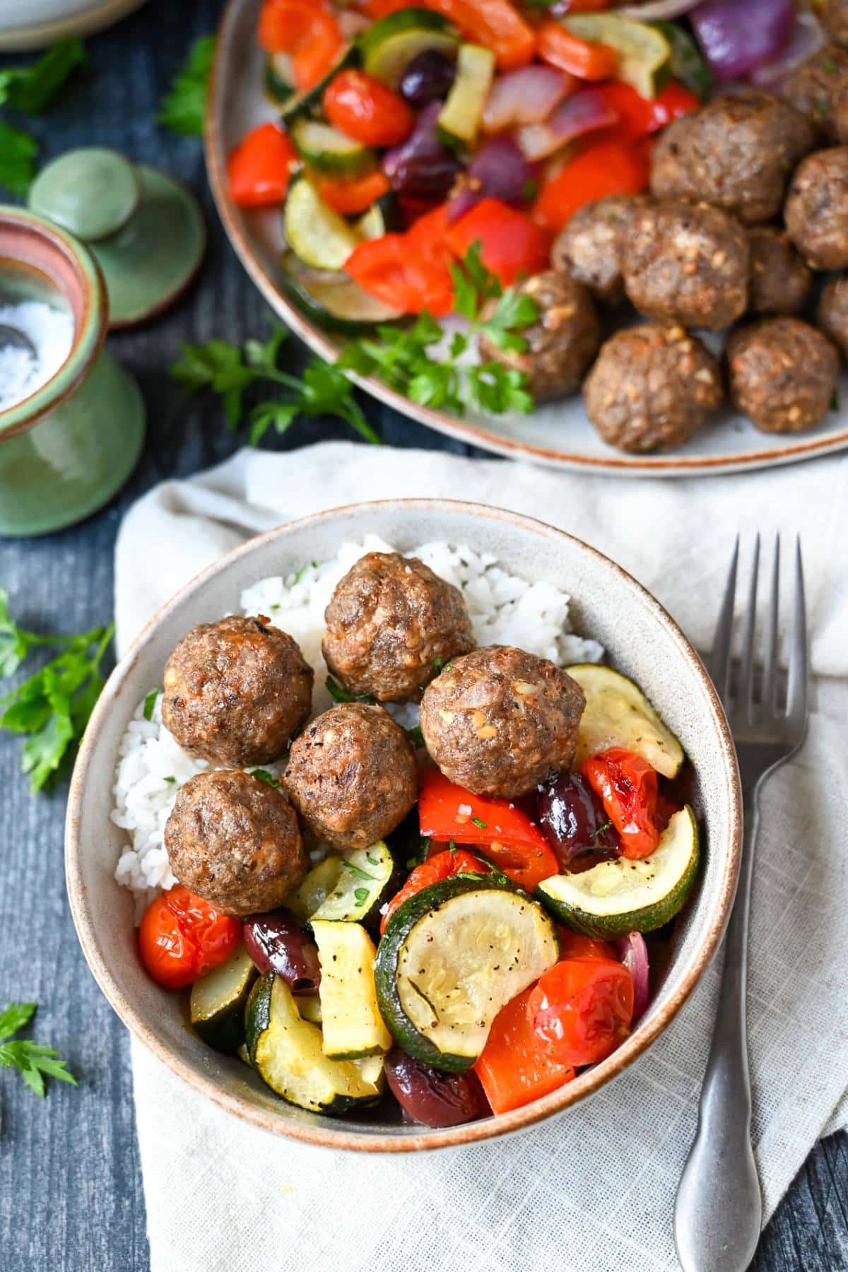 Greek meatballs in a bowl with rice and vegetables with a napkin, fork, and a plate of meatballs and veggies