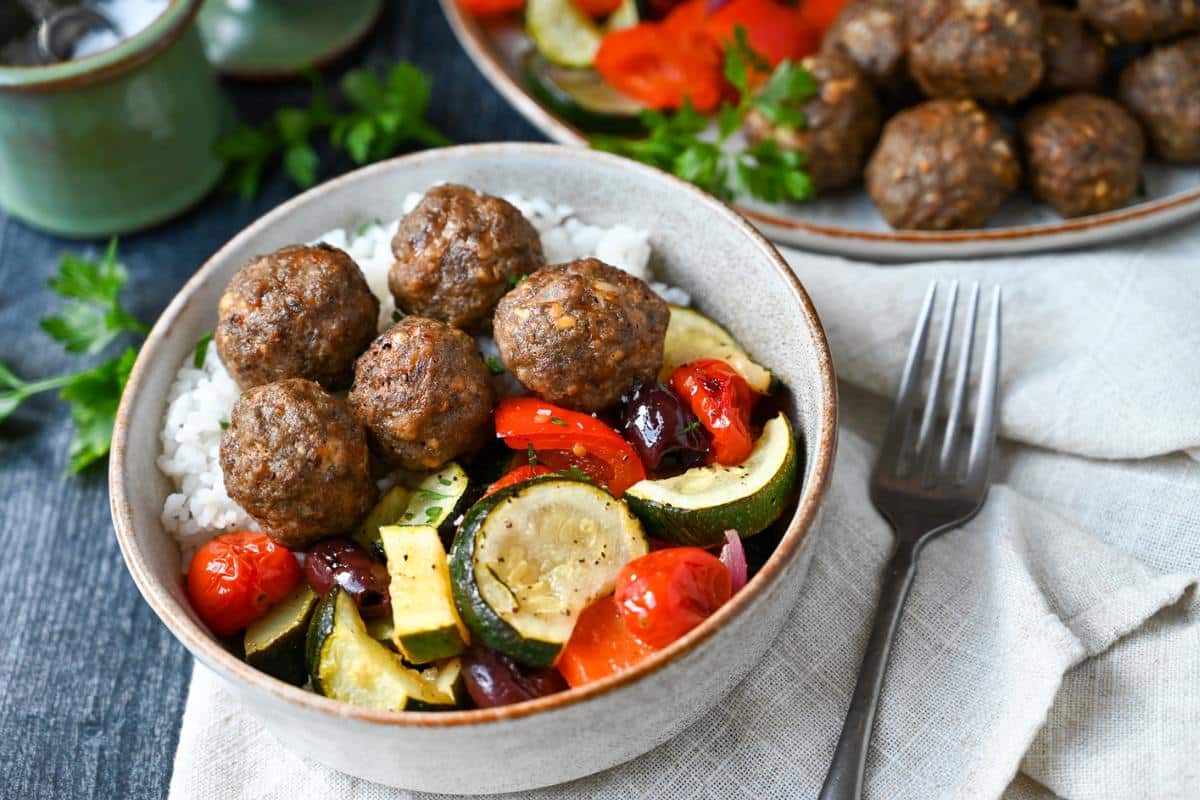 meatballs in a bowl with rice and roasted vegetables with a napkin and fork next to it