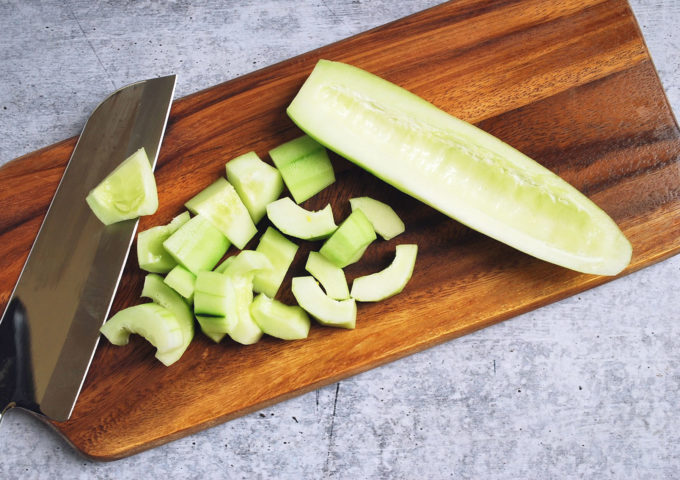 A knife sitting on top of a wooden cutting board with a seeded and sliced cucumber