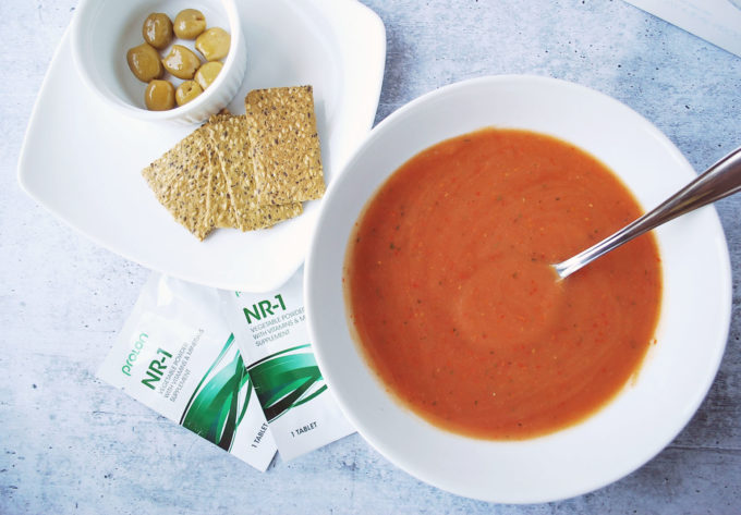 A bowl of soup and a spoon on a table, with crackers, olives, and packages of vitamins