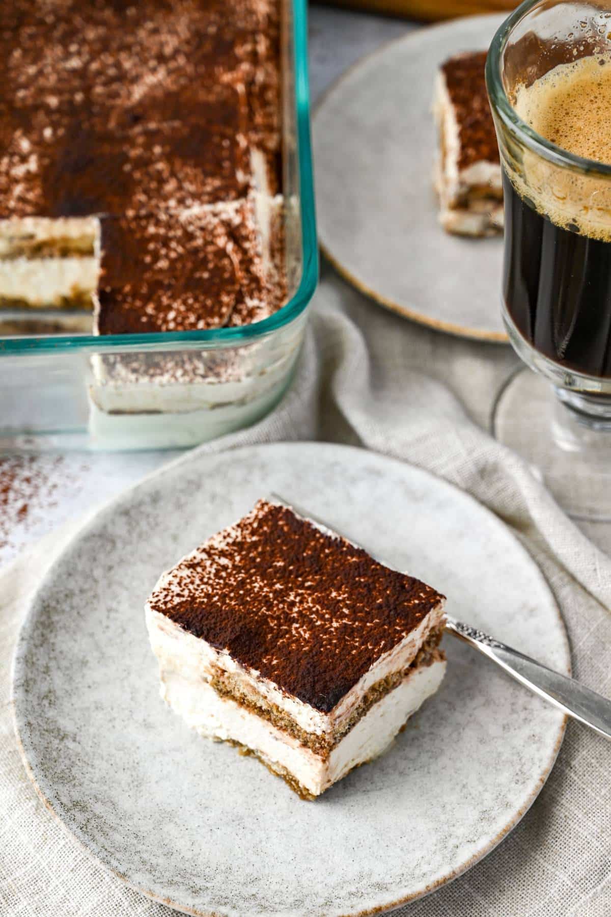 a slice of tiramisu on a plate with a fork and a cup of coffee behind it