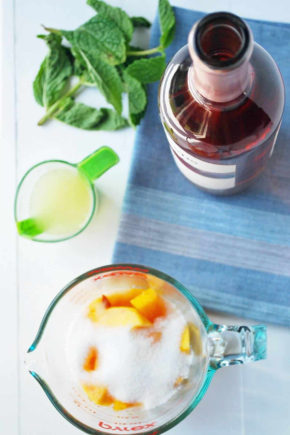a bottle of rose with fresh mint, lemon juice, and a measuring cup with peaches and sugar