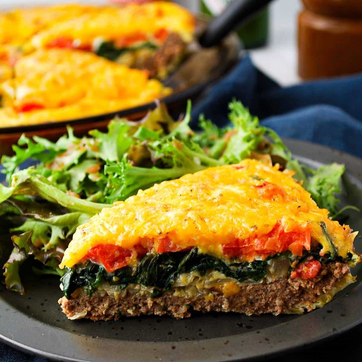 slice of cheeseburger quiche on a plate with salad with a quiche in the background