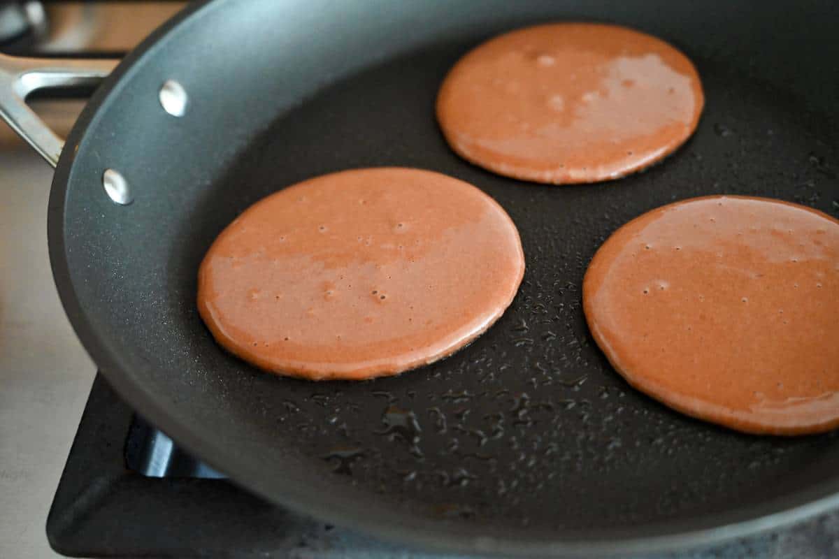 three chocolate pancakes cooking in a skillet ready to flip