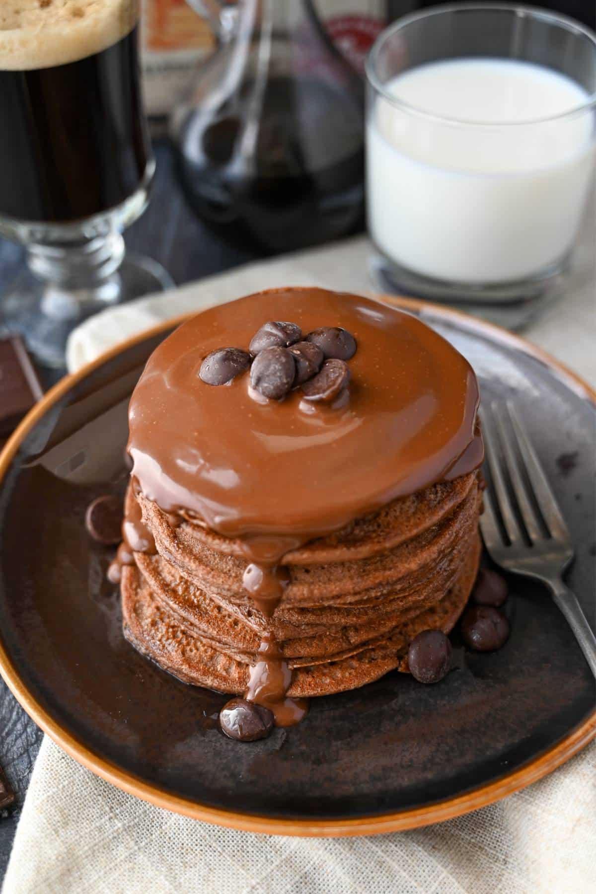 a stack of chocolate pancakes with poured healthier chocolate sauce and a glass of milk and coffee in the background