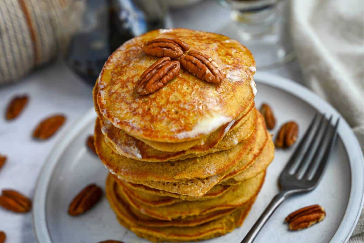 side view of a stack of pancakes with pecans and maple syrup with a cup of coffee behind it