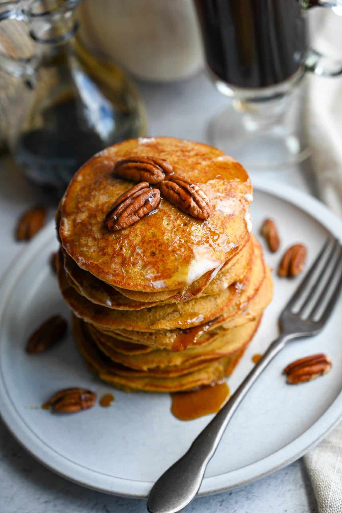 a large stack of pumpkin oatmeal pancakes topped with pecans and maple syrup
