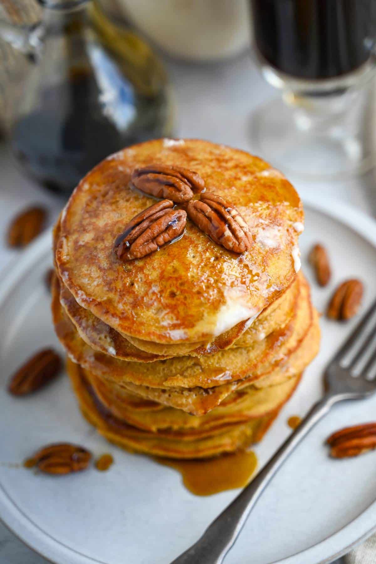 stacked pumpkin pancakes with a jar of maple syrup, a cup of coffee and pecans sprinkled around it
