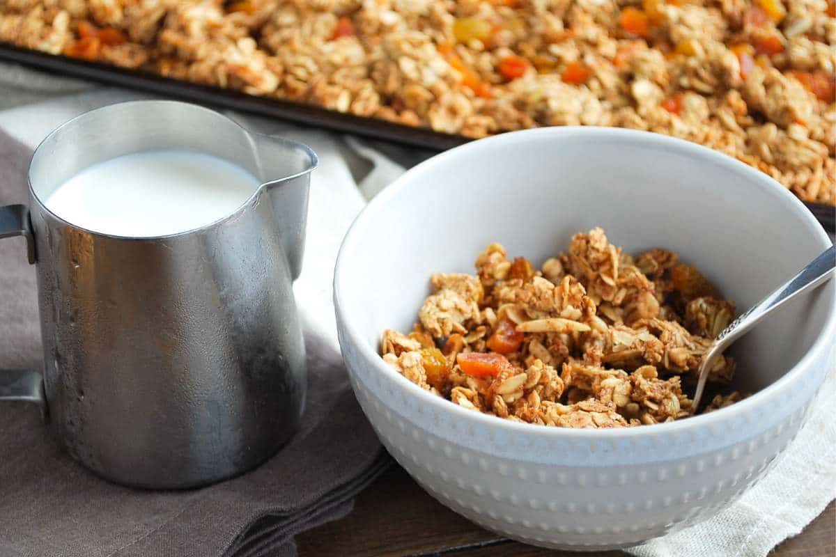 a bowl of vanilla almond granola with a carafe of milk and pan of granola in the background