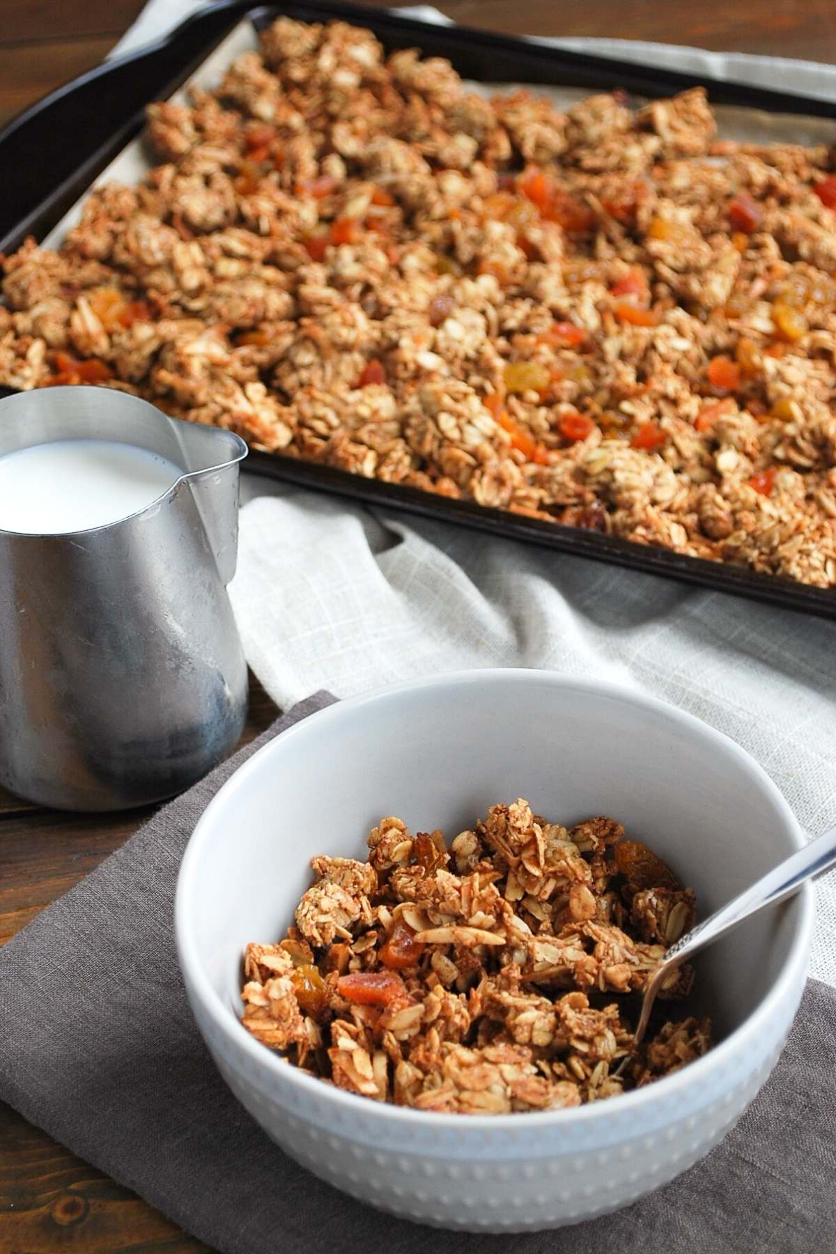 a bowl of granola with a carafe of milk with pan of granola in the background