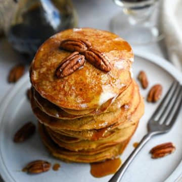 close up of a stack of pumpkin pancakes with pecans and maple syrup on top with a fork next to it