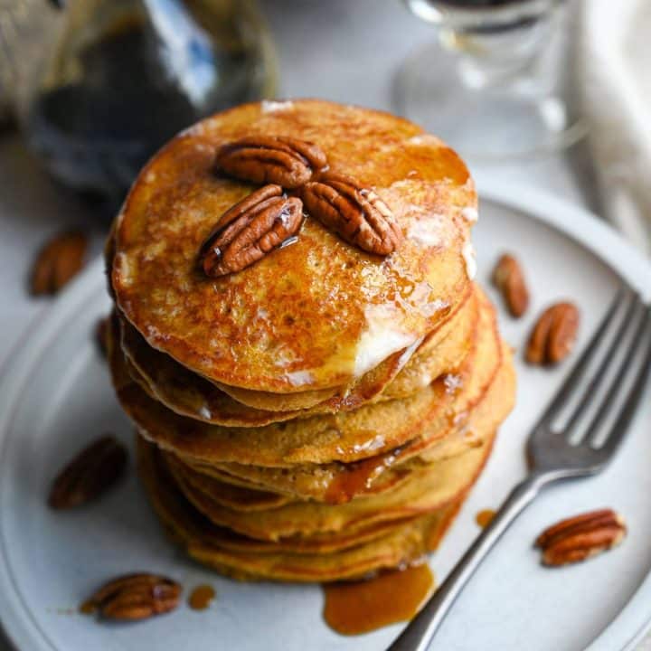 close up of a stack of pumpkin pancakes with pecans and maple syrup on top with a fork next to it