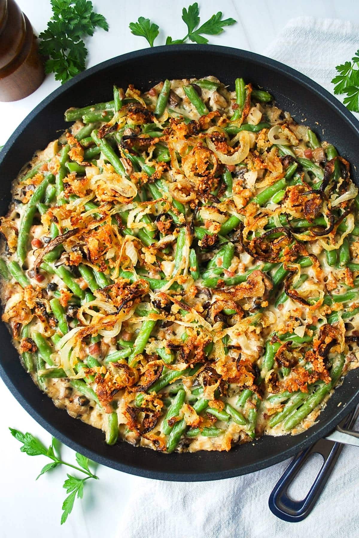 a skillet with loaded green bean casserole with a napkin, serving spoon, and fresh parsley sprinkled around the skillet