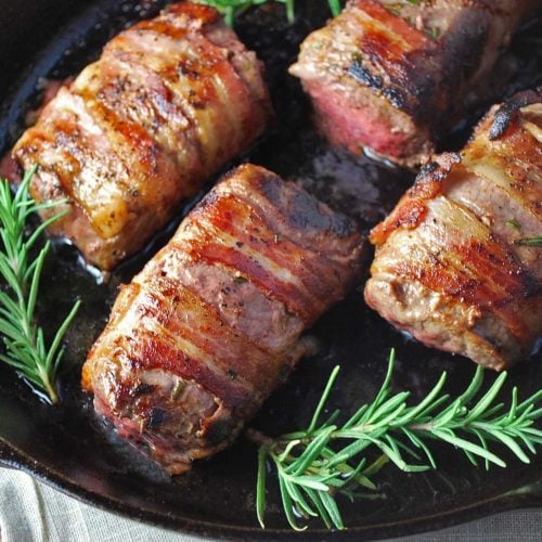 venison tenderloins wrapped in bacon seared in a cast iron skillet with fresh rosemary sprigs