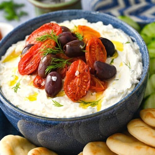 close up of a bowl of whipped feta dip topped with fresh dill, roasted cherry tomatoes, and olives
