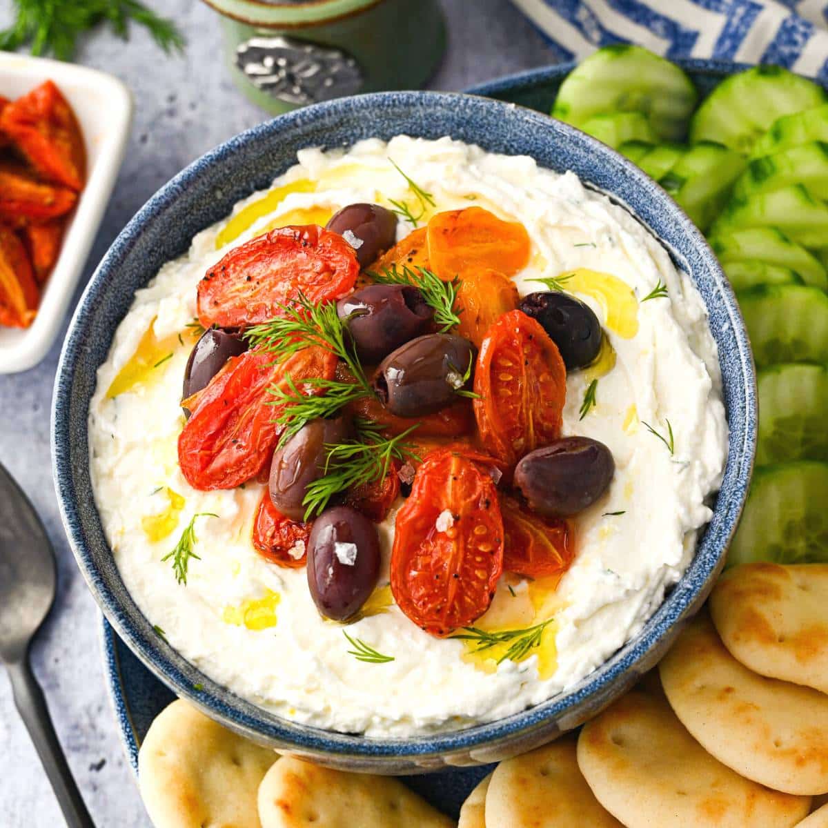 whipped feta dip in a blue serving bowl with a side of roasted tomatoes, cucumbers, and mini Naan bread rounds