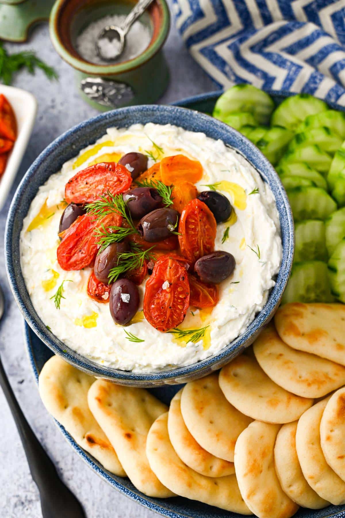 a bowl of whipped feta dip topped with roasted tomatoes and kalamata olives drizzled in olive oil with Naan bread and cucumbers on a platter