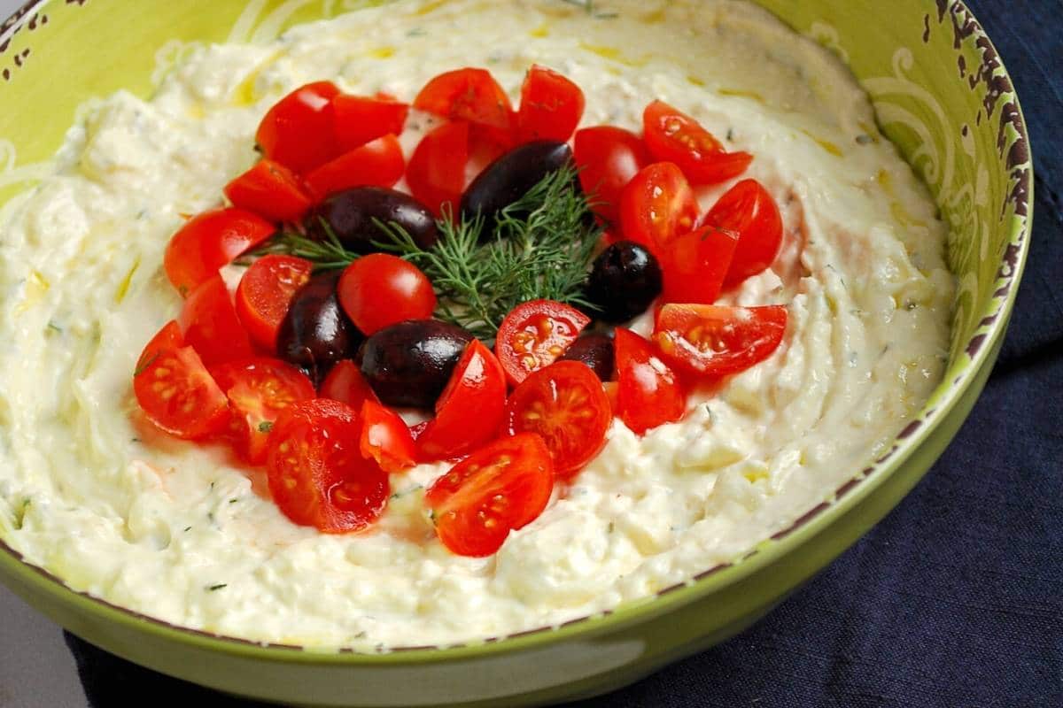 close up of a bowl of Greek feta protein dip topped with chopped tomatoes, fresh dill, and olives
