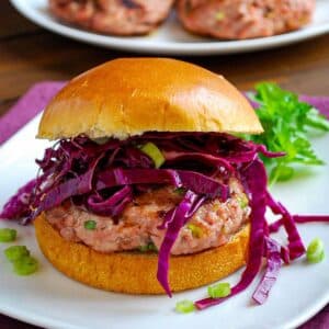 close up of a grilled asian pork burger with red slaw and a brioche bun on a plate with greens