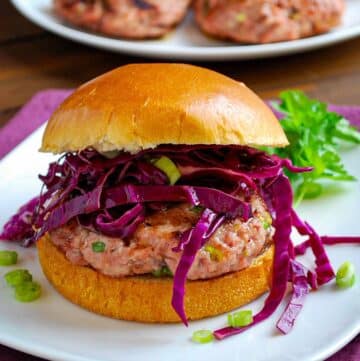 close up of a grilled asian pork burger with red slaw and a brioche bun on a plate with greens