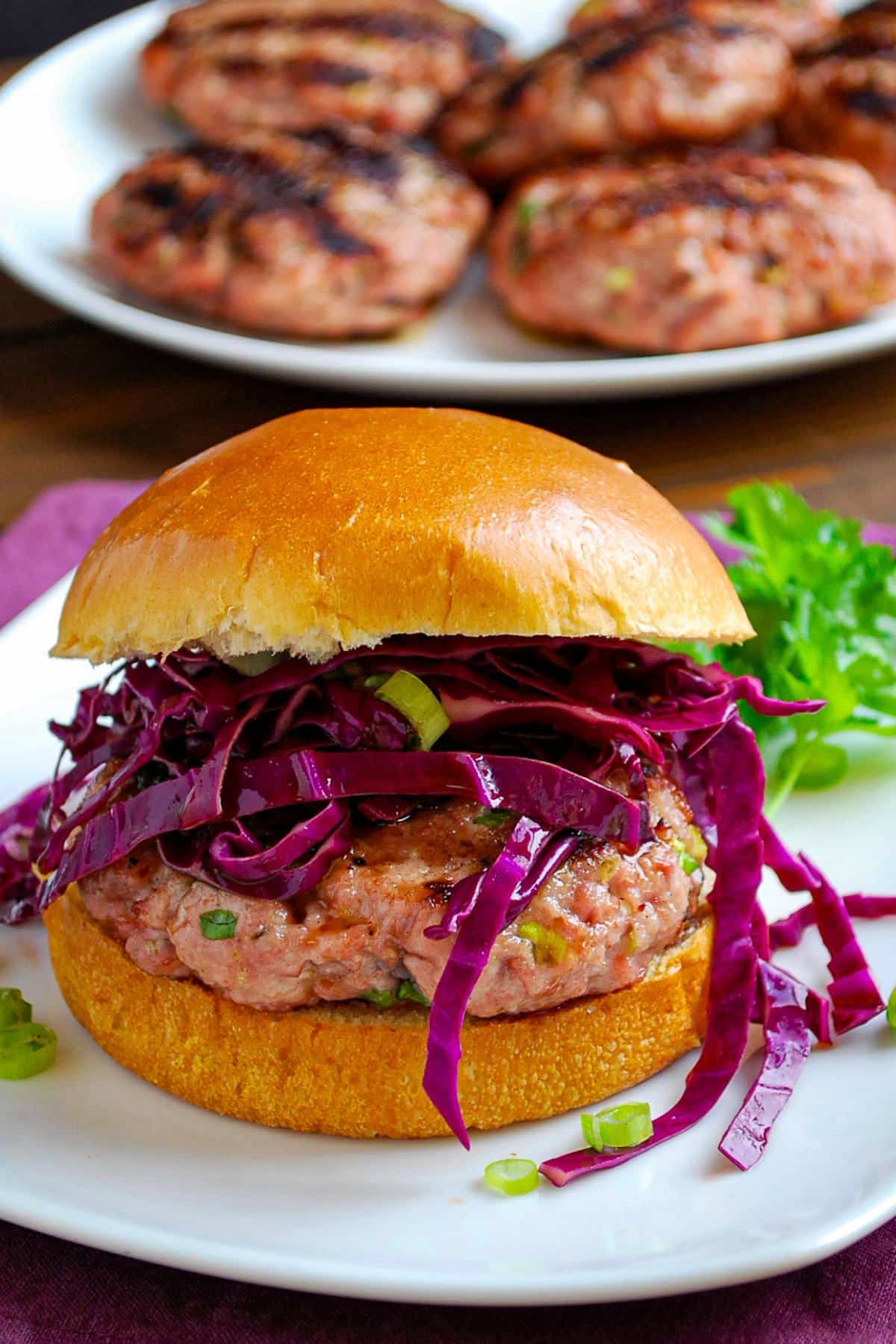a grilled pork burger topped with red cabbage slaw on a plate with a plate of burgers behind it