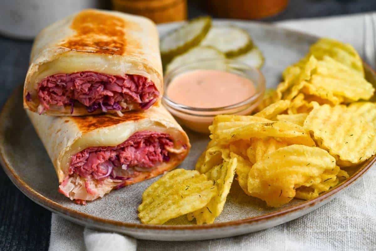 a sliced reuben on plate with a dish of dressing, chips, and a pickle