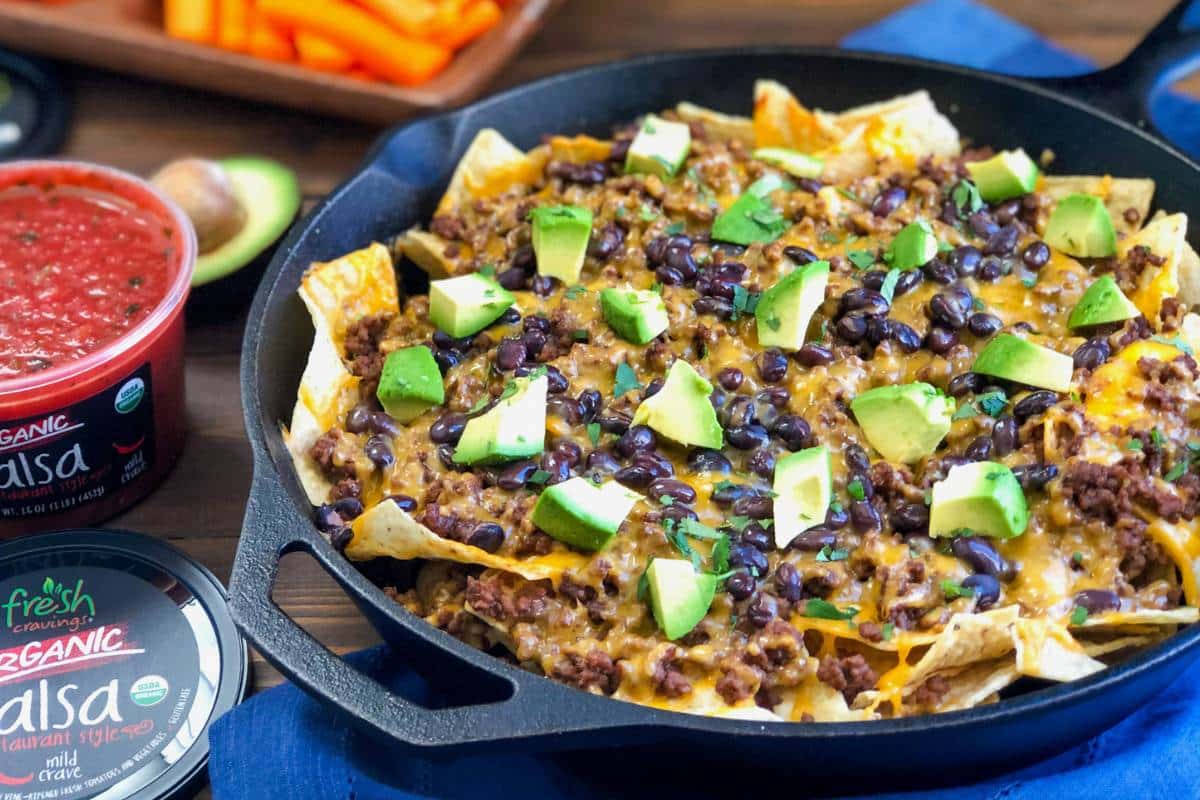 a cast iron skillet with cooked beef nachos with a tub of salsa and veggies in the background