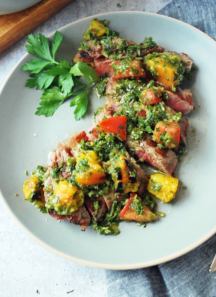 plate of sliced strip steak topped with peach and tomato chimichurri sauce with sprig of parsley