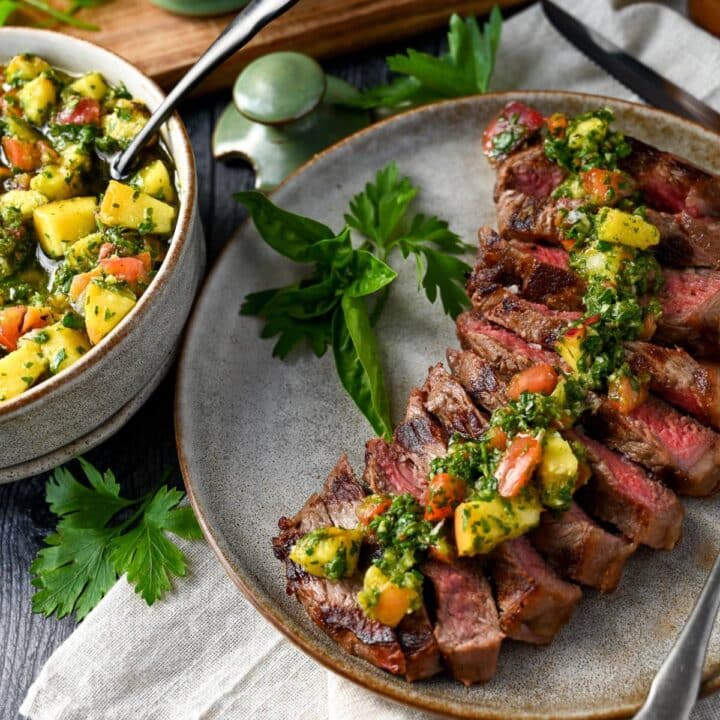 grilled steak sliced on a plate with chimichurri sauce and fresh herbs with fresh herbs around the plate