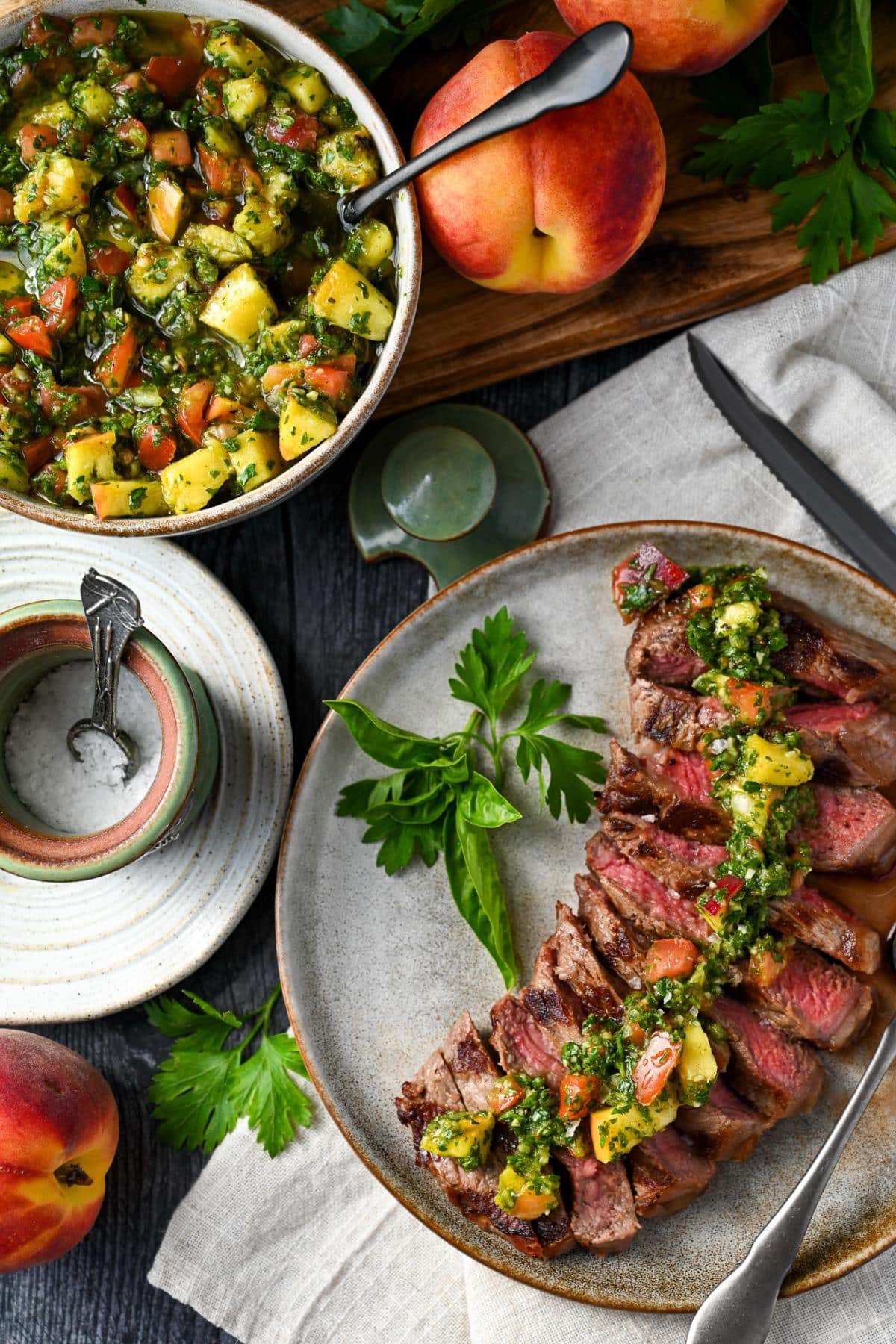 a sliced steak with peach chimichurri on top with fresh herbs on the plate and peaches around it with a bowl of chimichurri 