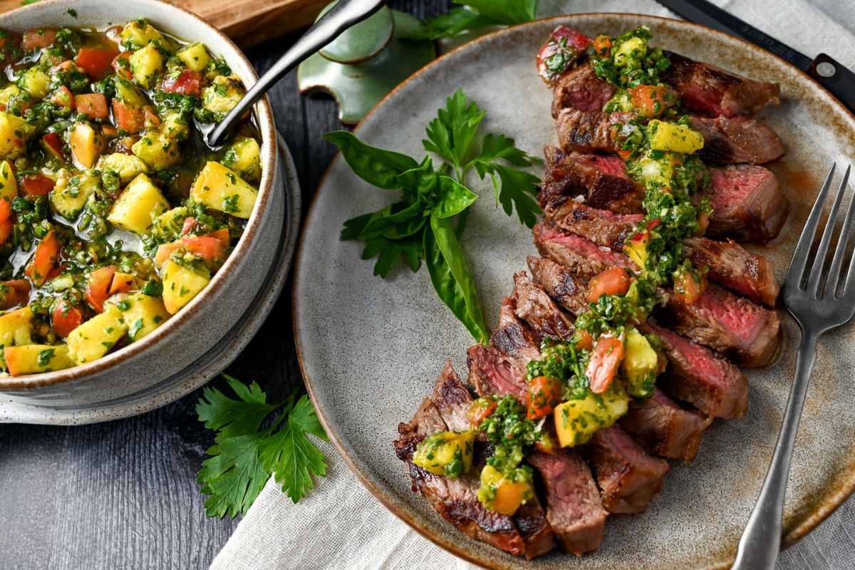 a sliced new york strip on a plate with grilled peach chimichurri sauce drizzled on top with a bowl of chimichurri next to it