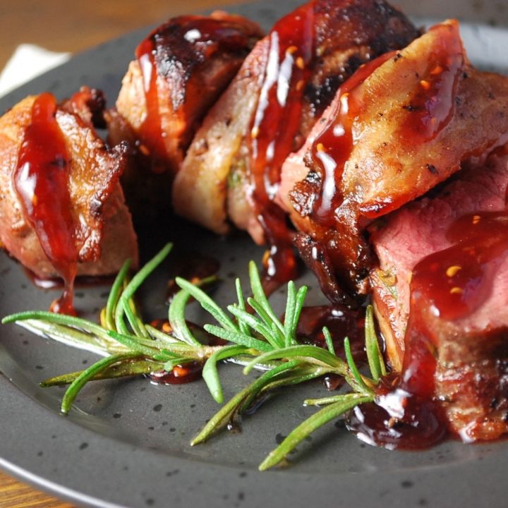 sliced bacon-wrapped Venison backstrap on a plate with rosemary and raspberry sauce