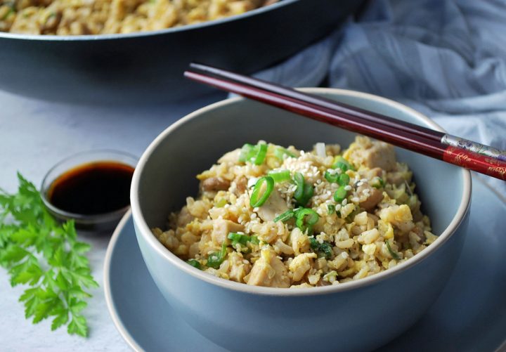 fried rice in a grey bowl with green onions and sesame seeds on top with chopsticks laying on top of the bowl and sauce and a skillet in the background