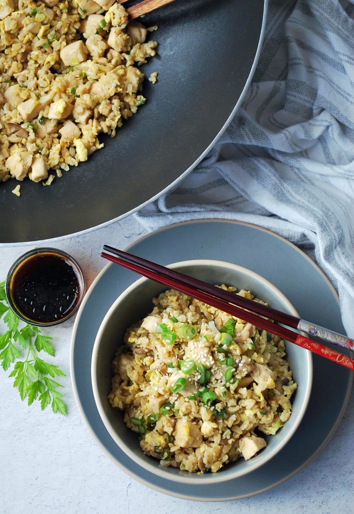 overhead photo of a skillet of fried rice with a bowl of fried rice with chopsticks in the front. There is also a dish of soy sauce and a sprig of parsley on the side