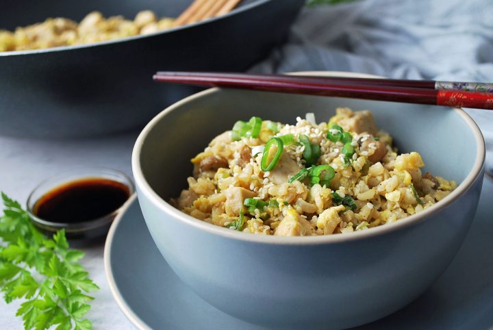 Close up photo of fried rice in a grey bowl with chopsticks laying on top and parsley and bowl of sauce in the background