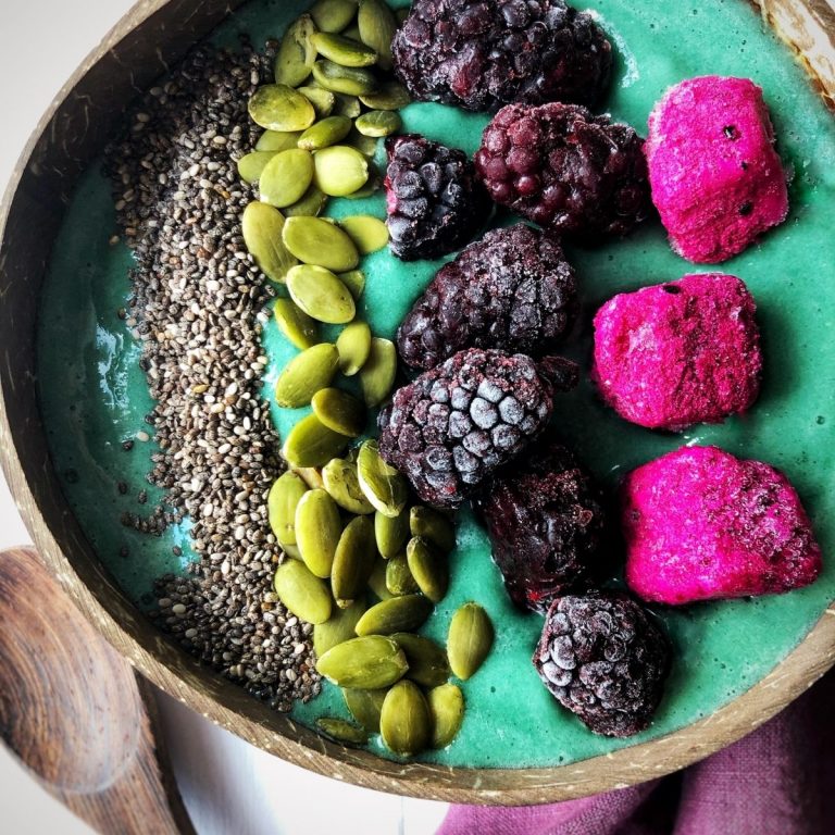 a close up photo of a blue smoothie in a coconut shell topped with frozen fruit and seeds